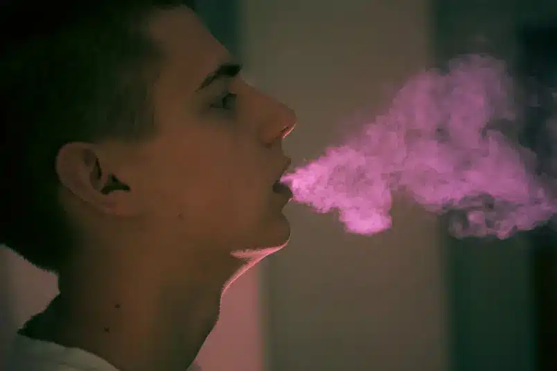 A teenage boy blows smoke out of his mouth with a pink light coming from the background