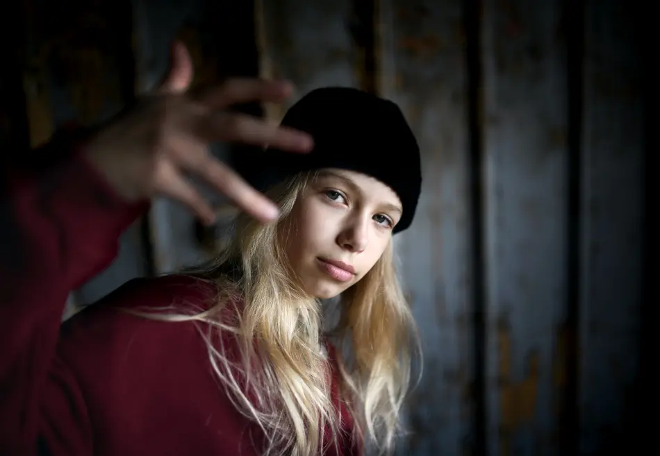 A blonde teenage girl with a beanie looking at the camera and waving