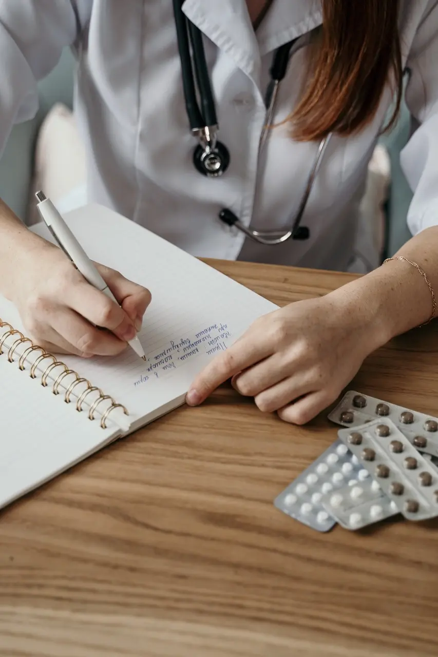 Doctor in a white coat writing down a Benzodiazepine prescription on a notepad, pills are next to her on the table