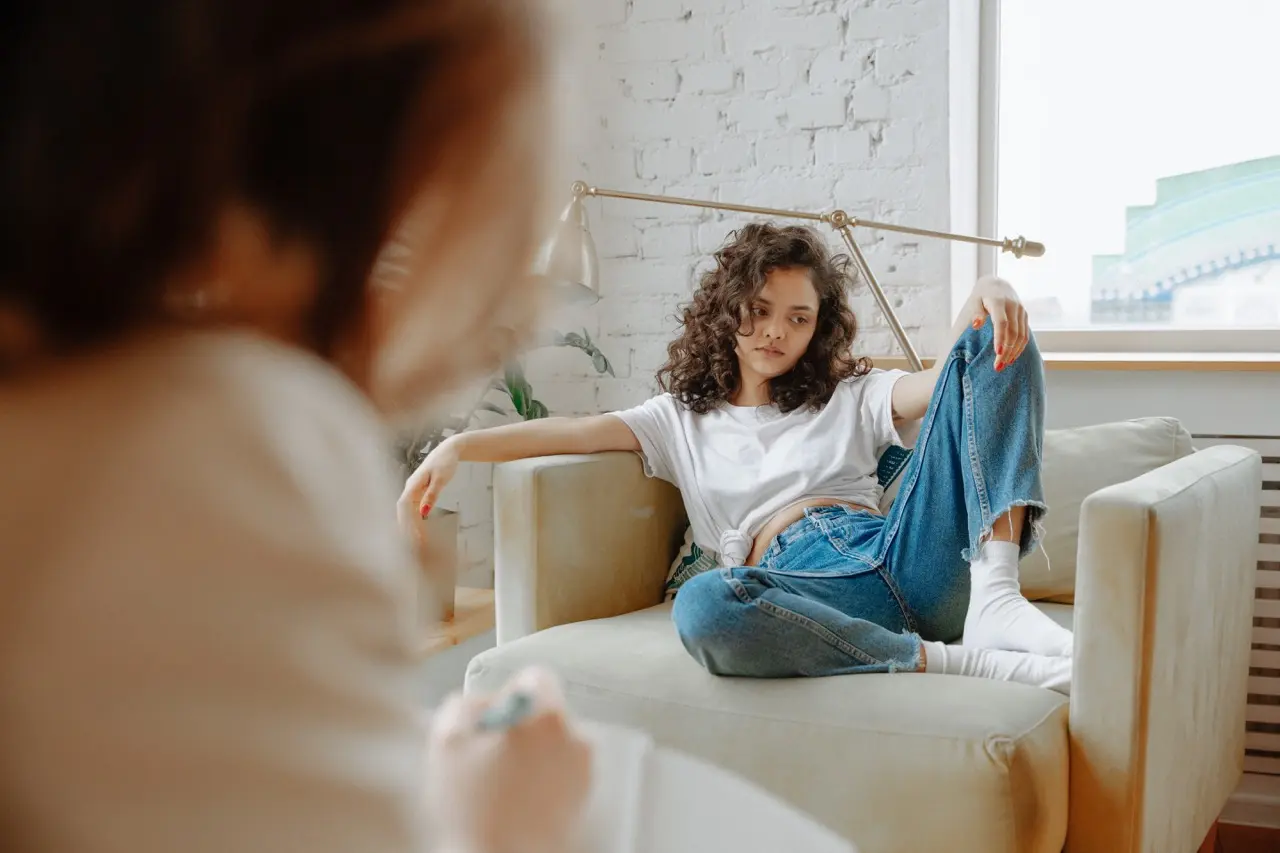 Curly haired woman wearing a white shirt and jeans sits on a white couch resting her arm on her raised knee during a stimulant treatment 1-on-1 therapy session, looking anxiously to the distance