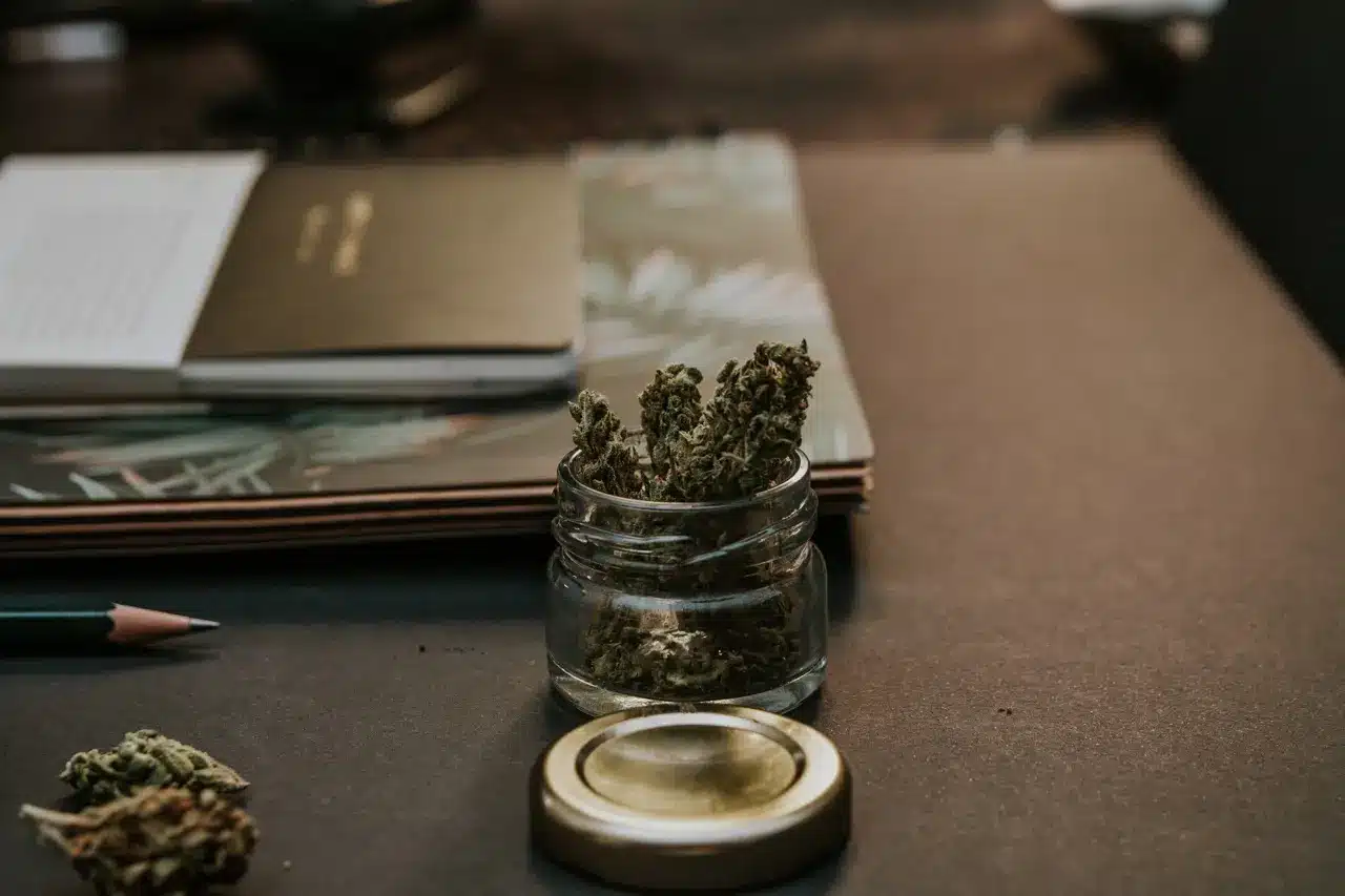 A jar of dried marijuana sitting on a wooden desk with notepads next to it