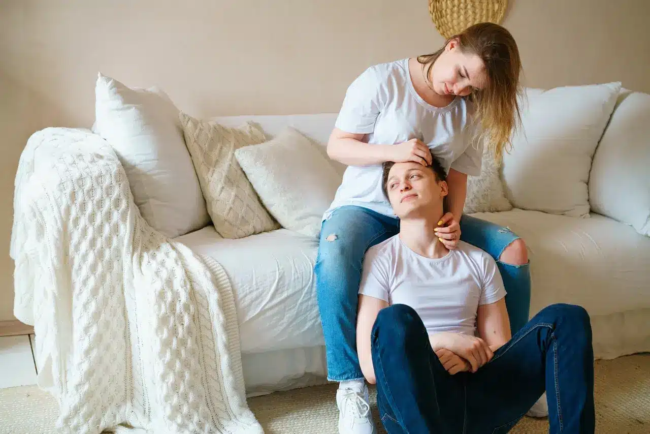 Man suffering from stimulant addiction sitting on the floor looking anxious with his head leaning on a woman sitting on a white couch behind him, holding his head in comfort
