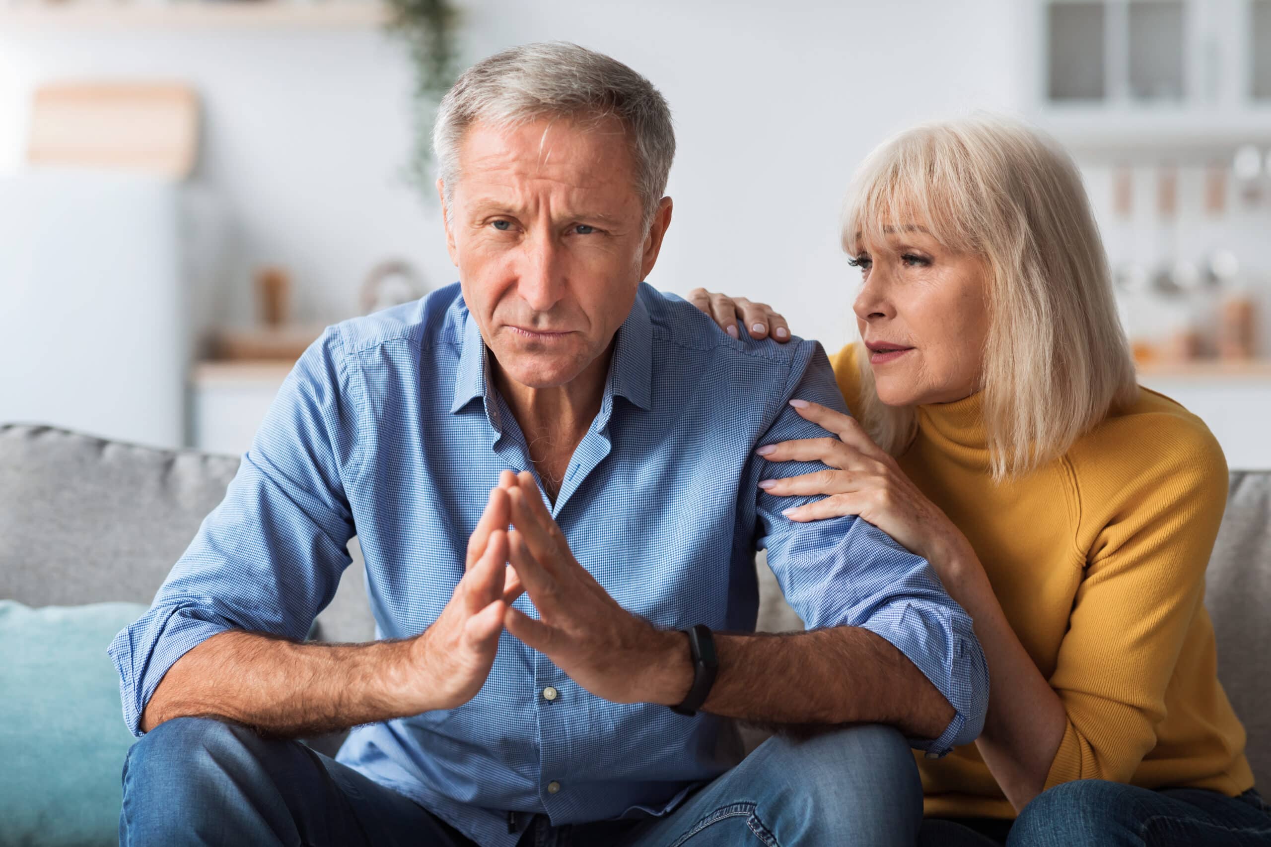 Worried Senior Wife Talking To Concerned Husband Hugging Him, Having Problems In Married Life Sitting On Couch At Home. Woman Comforting And Supporting Man During Crisis Indoor