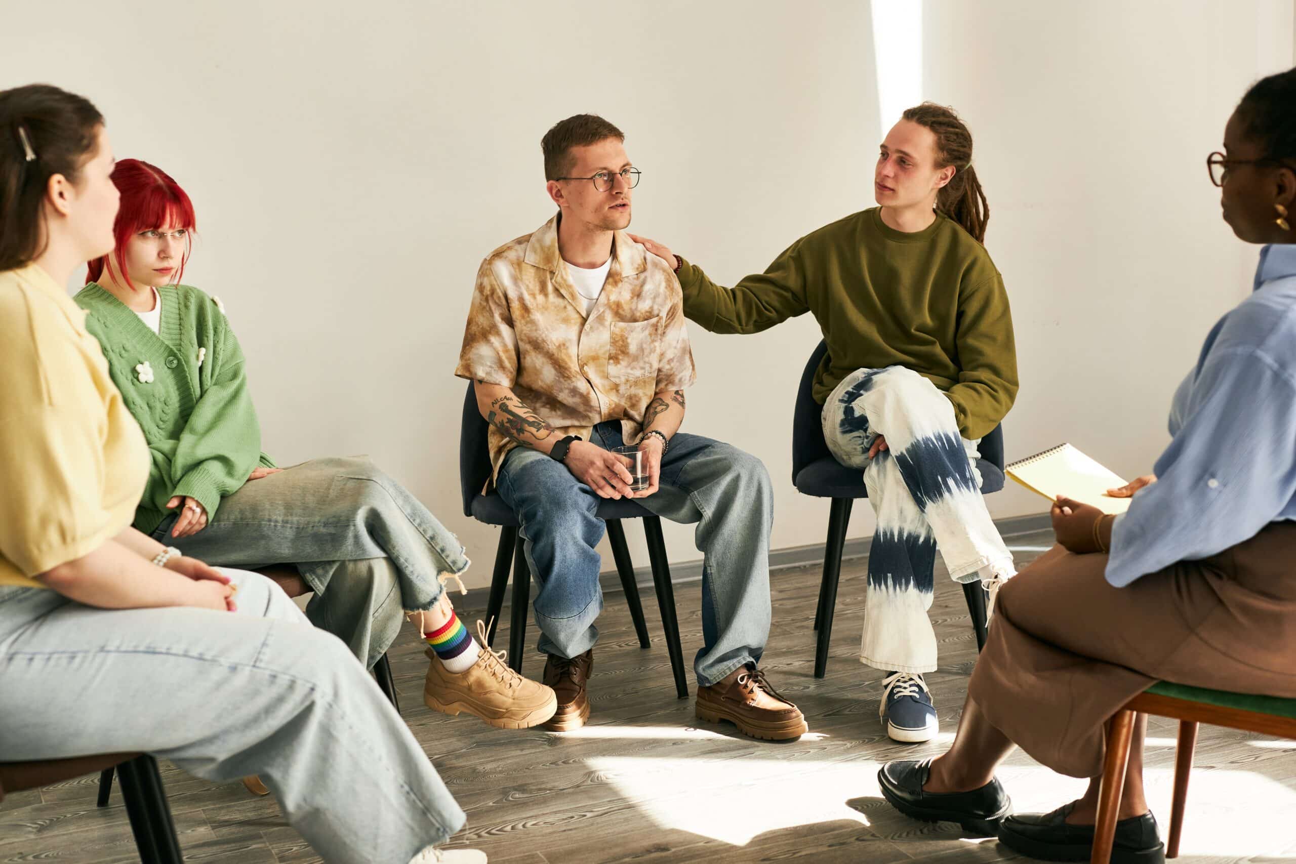 Diverse group of individuals engaging in group therapy session, listening and participating in an informal setting with bright flooring and relaxed atmosphere